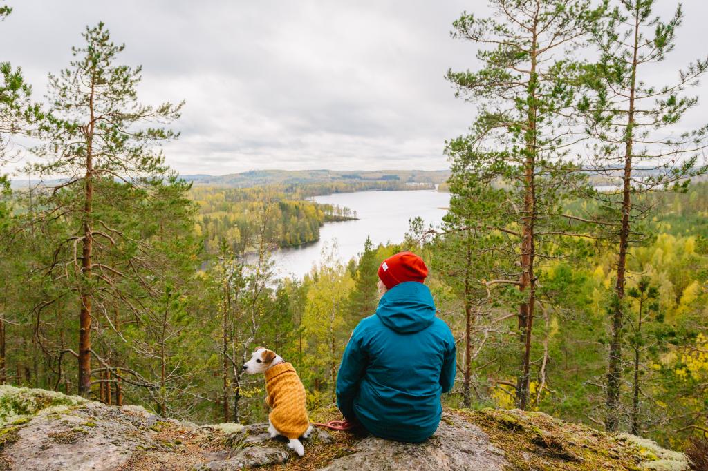 Nainen istuu koiran kanssa korkean kallion laella ja katselee alas näkymiä sokkeloiseen saaristoon.